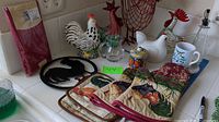 Full view of assortment on kitchen counter, showing various rooster and chicken themed kitchen decor items including pot holders, figurines, mugs, and a metal rooster trivet.