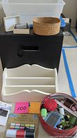 Several storage containers including plastic bin, wooden shelf organizer, and round red container with sewing items