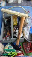 Blue plastic storage bin with various tools inside including hammer, scrapers, scissors, wire cutters, hand tools, and assorted packs.