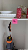 Photo showing white dishes on upper shelf and Oreck vacuum cleaner with orange and yellow accents on lower shelf with decorative red candles.