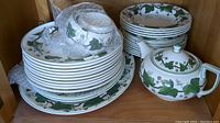 Stack of Wedgwood ivy pattern dinner plates, smaller plates, bowls, and teapot inside cabinet shelf.