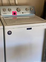 Front view of white Maytag top load washing machine showing control panel with dials and machine body with Maytag logo.