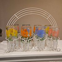 Front view of eight floral painted glasses in a white metal caddy, showing colorful flower designs on each glass and floral shapes on the caddy holders