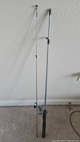 Full view of Shakespeare fishing rod and reel combo standing upright against the wall on carpet.