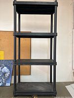 Black plastic shelving unit with four tiers, shown against a wall and corkboard.