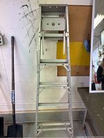 Front view of silver metal step ladder showing multiple steps and a small platform at the top, positioned against a white wall next to a shovel.