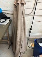 Photo showing beige folded umbrella leaning against a wall next to a table, with a black metal pole nearby.