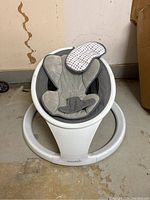 Front view of the Munchkin baby swing showing the white circular frame and gray cushioned sling seat with attached polka dot padded headrest.