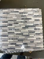 Pack of mosaic backsplash tiles showing the varied gray and white linear strips.