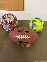 Photo showing three sports balls: Adidas soccer ball with red, white and black star design, yellow and blue handball, Wilson branded brown football