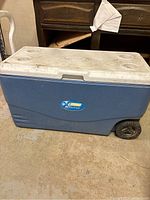 Closed view of blue and white Coleman cooler showing wheels, lid with cup holders and logo.
