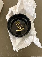 Top down view of black ceramic ashtray showing gold OVO owl logo on the interior