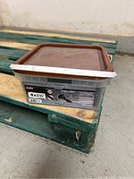 Plastic bucket with brown lid labeled 8x1 1/4 in black floor screws placed on a green wooden pallet.