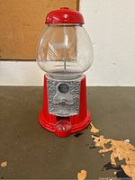 Red vintage-style gumball coin machine on flat surface showing front and top with clear round globe and silver coin slot.
