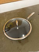 Hexclad frying pan with glass lid on a brown surface, showing stainless steel handle and glass lid with metal rim.