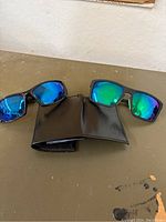 Two pairs of black rectangular frame sunglasses with reflective blue-green lenses next to a black bifold wallet on a textured brown surface