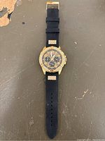 Full view of the men's Guess watch showing the black strap, gold-tone case with decorative stones, multi-dial face, and stone accents on the strap.