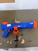 Blue and orange Nerf Dog Shooter toy designed for dogs, placed on a worn tabletop, showing overall condition and details such as trigger and barrel.