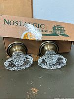 Pair of clear faceted glass Nostalgic door knobs mounted on brass bases, displayed in front of original Nostalgic House cardboard box.