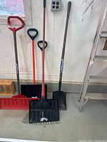 Three snow shovels, one rake, and one gardening shovel lined up against a wall, showing condition and full length of handles and blades.