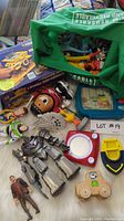 Overview photo showing various toys including action figures, a boxed toy set, an Etch A Sketch, and a green bag with toy pieces.