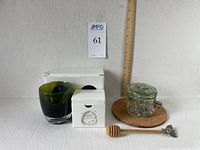 Lot displayed with white background, showing boxed beeswax tealight candles, green and black Glassybaby glass candle holder, glass honey jar with embossed honeycomb pattern and metal bee details, wooden honey dipper with bee ornament, sitting on cork coaster.