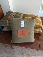 Five large throw pillows stacked on a wooden surface. The center pillow is a Ralph Lauren pillow in tan fabric with brown leather corner accents and a leather label patch.