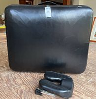 Closed black portable massage table case with handle and detached black face cradle on wood floor.