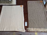 Two blankets side by side on a table. Left is a cream cable knit Ralph Lauren throw with visible label, right is a beige woven Sunbrella blanket with fringes and an orange size sticker.