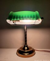 Full view of the lit vintage banker’s lamp showing green glass shade and brass base on a gray background.