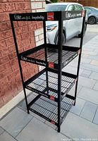 Full view of black metal vintage Coca-Cola display rack with four wire shelves and branded logos on shelf edges and sides.