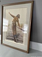 Full view of the framed lithograph depicting a person balancing on a railroad track, showing the frame and surrounding wall.