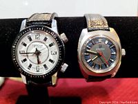 Pair of wristwatches displayed on a stand: left is Lucerne Alarm watch with black bezel and white dial; right is Accutron watch with blue and gray dial and orange details, black leather straps.