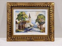 Framed oil on board painting showing village street, church steeple, trees, and figures.