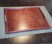 Wooden tray with brass handles on woven placemat, visible rich wood grain and polished finish.