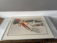 Framed skiing lithograph showing a skier in red and yellow passing flags with crowd in background.