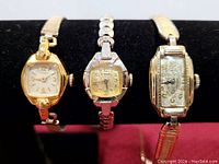 Three vintage wristwatches displayed on a black stand showing their faces and bands.