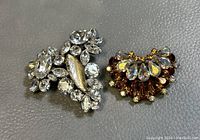 Two vintage Sherman signed crystal brooches side by side on a gray surface, showing clear crystals and a distinctive silver leaf detail on one and amber and clear crystals on the other.
