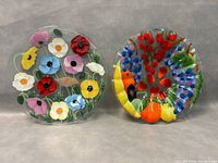 Two round fused glass decorative plates side by side on a gray background, one with colorful flowers and one with a bird and berries design.