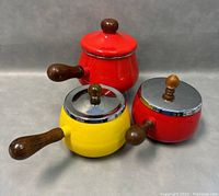 Three vintage enamel saucepans grouped together, two red and one yellow, with wooden handles and lids.