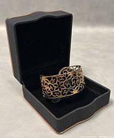 View of the gold tone pierced cuff bangle bracelet inside a black jewelry box, showing the intricate openwork floral and scroll pattern