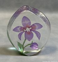 Front view of the crystal paperweight showing embedded purple flower and bud inside clear glass with textured edges.