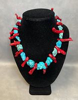 Front view of the necklace showing the alternating chunky turquoise beads and red coral branch-shaped beads on a black display stand.