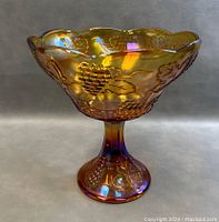 Side view of the marigold carnival glass compote bowl on pedestal, showing embossed grape and leaf design, scalloped rim, and iridescent finish.