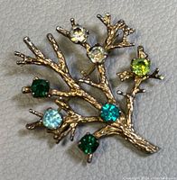 Front view of the vintage gold tone sterling silver tree branch brooch adorned with colored crystals on a textured light gray background.