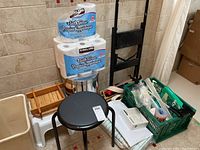 Overview of bathroom accessories lot showing bath tissue, stools, trash bin, trays, crate with supplies, and toilet step stool.