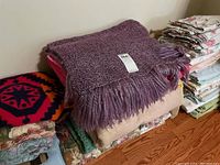 Stack of blankets and folded linens including purple fringed blanket and red/black patterned blanket on an oak floor.