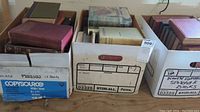 Three white boxes filled with various hardcover and vintage books, showing stacked titles with visible wear on bindings and covers.