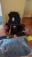 Full view of camping gear including backpack, sleeping bag in plastic, hatchet, folding chair, water bottles, and camping stove parts.