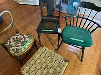 Three vintage chairs and one rush seat stool shown on wooden floor. Chair on left has floral upholstery with curved back, black chair in middle with damaged seat, black Windsor chair on right with green cushion, and rush seat stool in front.